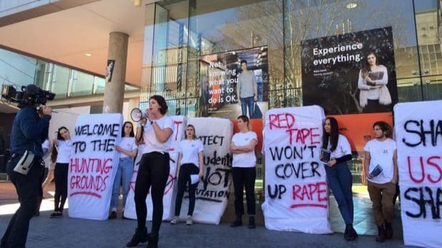 Members of the 2016 Women's Collective protesting outside the Law Annex building. They carry mattresses spray painted with slogans lie "Red tape won't cover up rape".