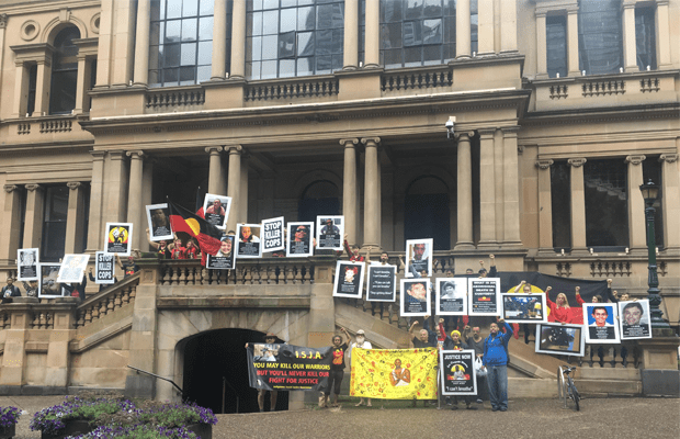 Protestors stand along the Sydney Town Hall steps, with images of deceased Indigenous people's faces.