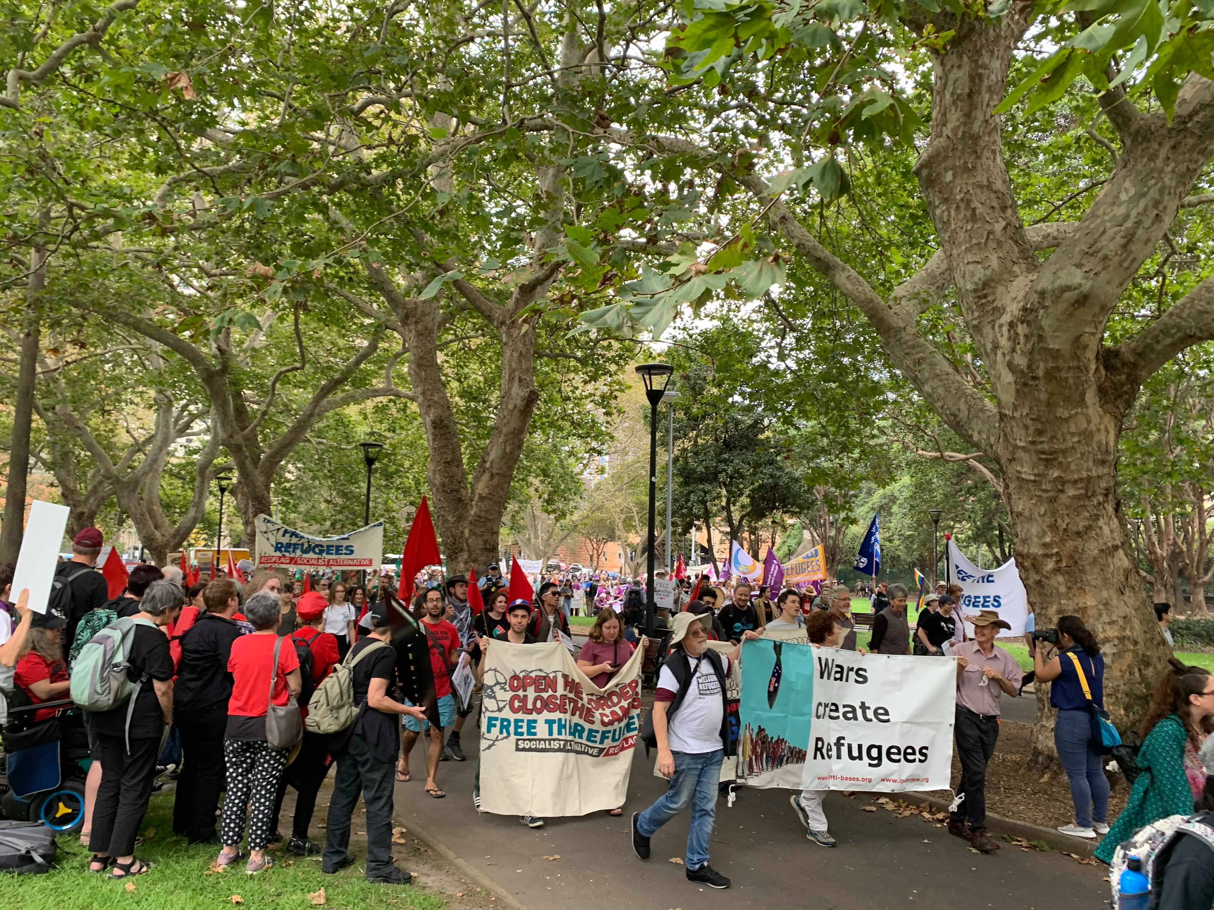 The contingent makes its way through Belmore Park, led by two banners: "Open the borders close the camps free the refugees now" and "wars create refugees" 