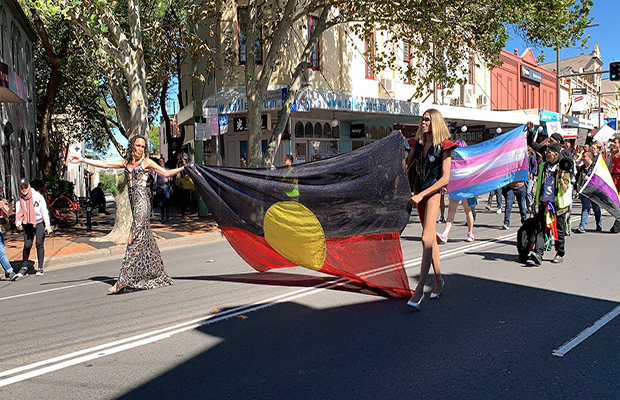 Two people walking along King street leading the rally holding an Aboriginal flag, followed by a Transgender Pride Flag