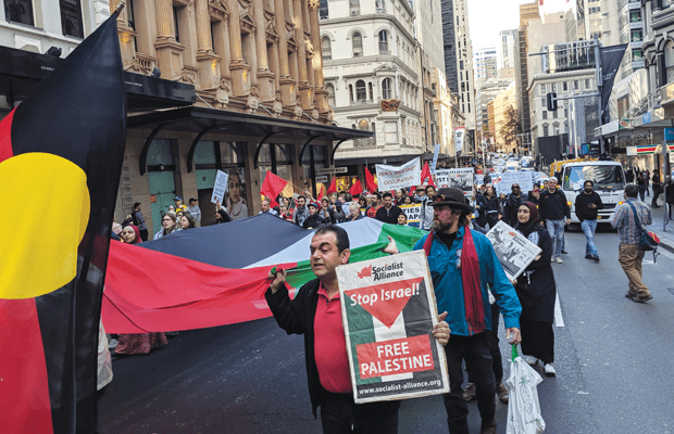 A man holding a Socialist Alliance paper with text "Stop Israel Free Palestine". Behind is a Palestinian flag being carried by many, and to the left is an Aboriginal flag.