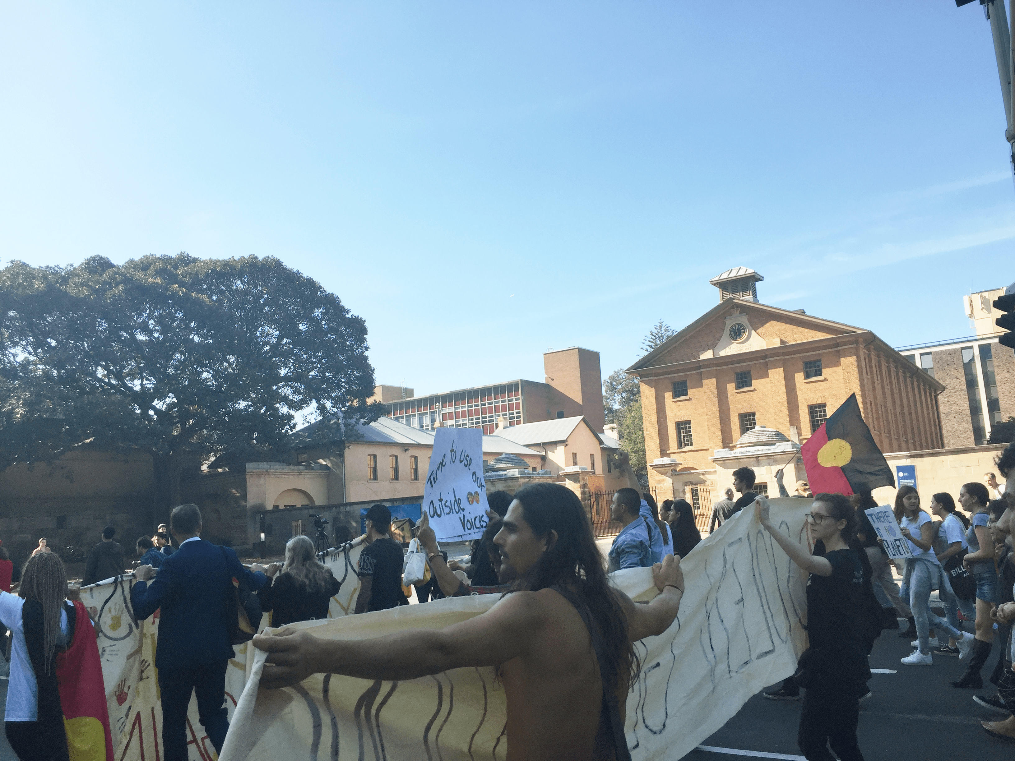 Photo of the march proceeding down Elizabeth Street towards the NSW Parliament House. A man stands shirtless holding a banner, and an Aboriginal flag flies in the background.