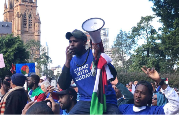 A photo of a protester hoisted on the shoulders of two others, holding a Sudanese flag and speaking into a megaphone