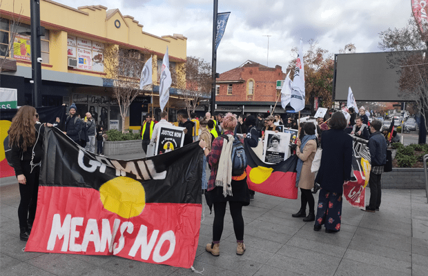 Photo: Protesters stand in Liverpool. A large Aboriginal flag is pictured, with white text that reads "GAMIL MEANS NO." There is a second Aboriginal flag, and other posters that read "JUSTICE NOW".