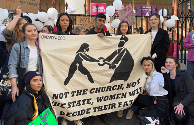 Photo of the University of Sydney Women's Collective, holding a banner which reads "Not the church, not the state, women will decide their fate", in front of NSW parliament house.