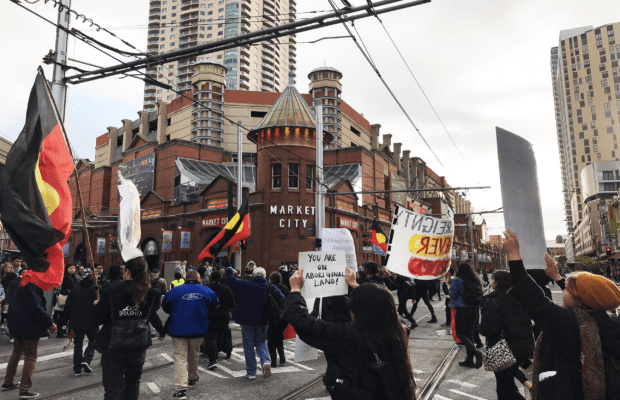 Protesters march through Market City intersection. Several Aboriginal flags are in the photo, which is taken from the midst of the crowd. In the middle is a sign that reads "You are on Aboriginal Land."