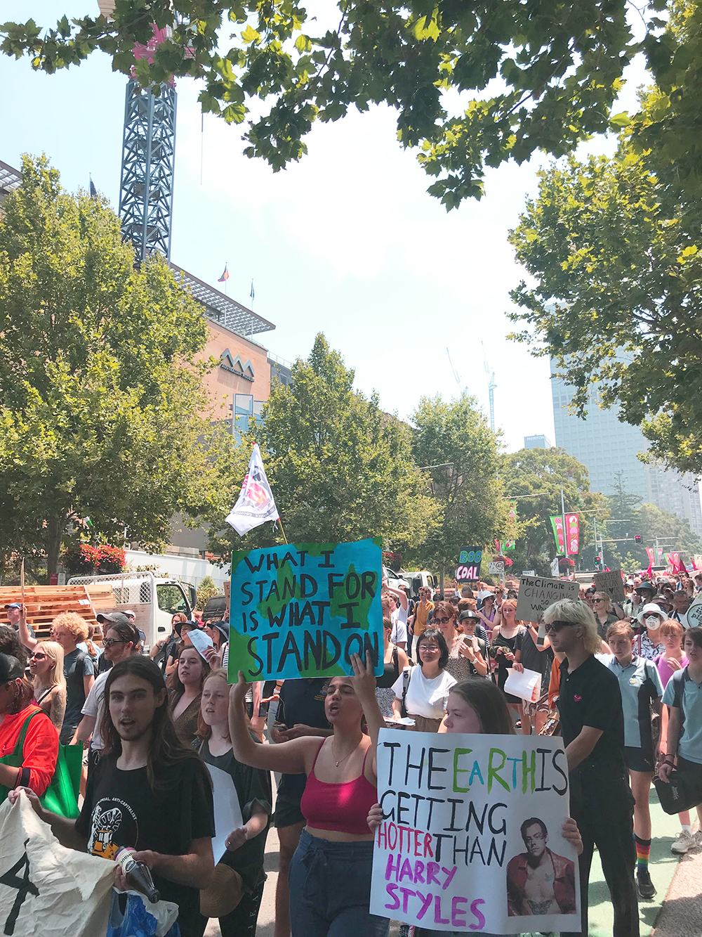 Protestors make their way along Williams Street, With two posters in shot. One reads "What I stand for is what I stand on" and another reads "The earth is getting hotter than Harry Styles." 