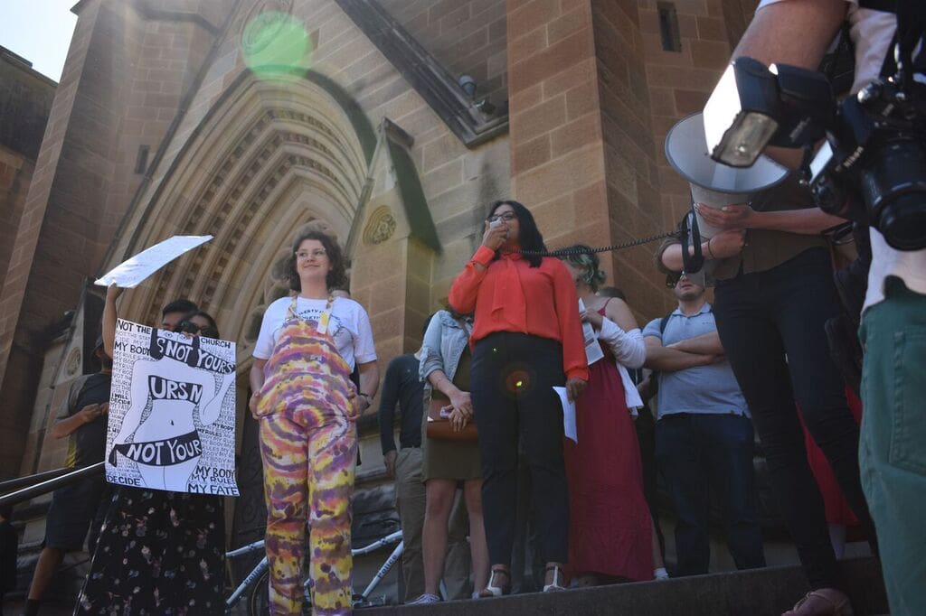 Greens MP Mehreen Feruqi standing at the top of St Mary's Cathedral's steps with a group of brightly coloured protestors, who are holding hand-painted signs and megaphones