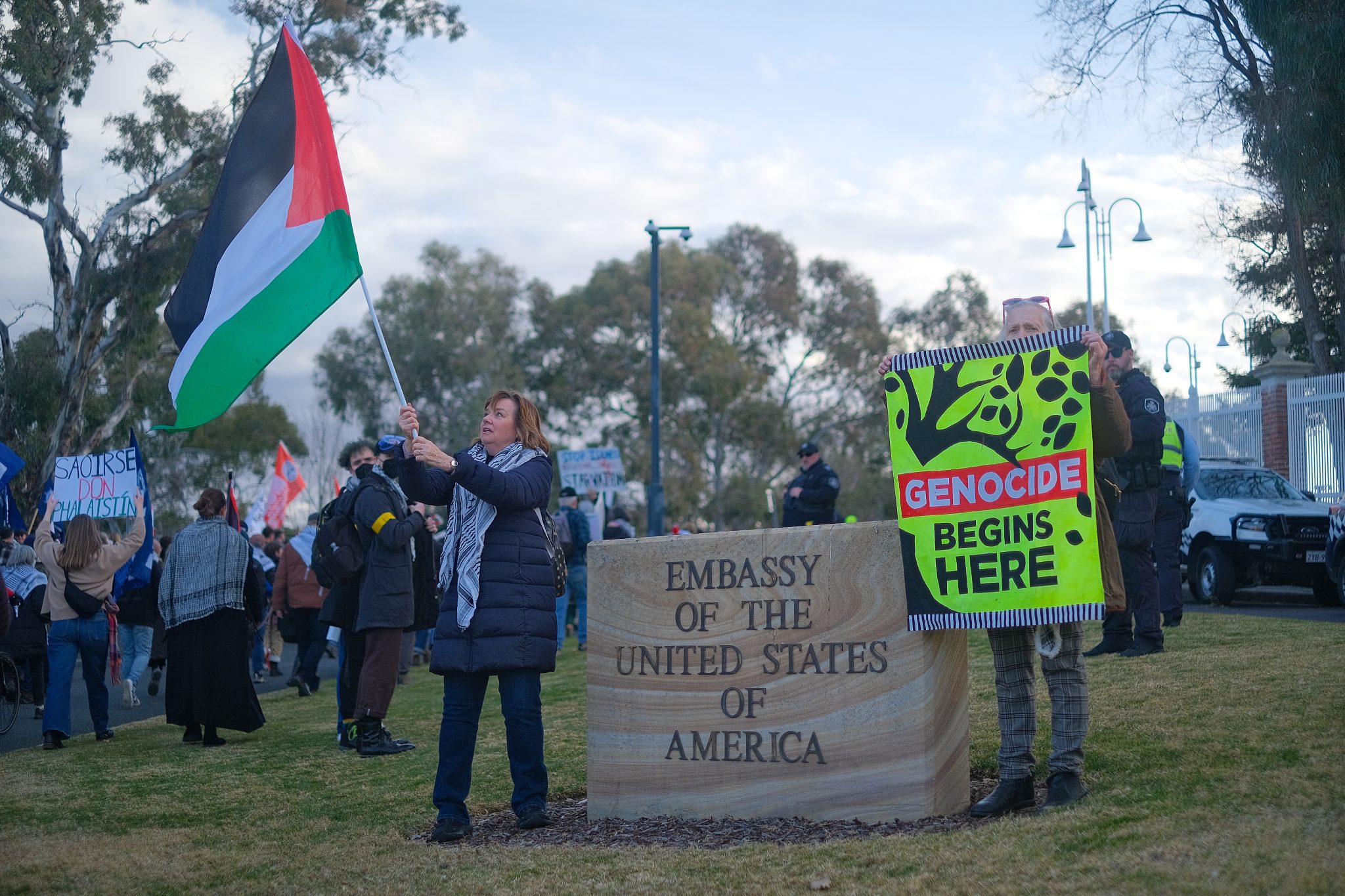 Protestors standing in front of a sign in front of the United States Embassy in Canberra. One protestor is holding the sign Genocide Begins Here.
